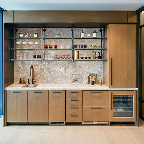 A well-equipped mini bar kitchen in the Library showcasing balanced contemporary interior design with warm wood and cool stone finishes. This beverage bar includes refrigerator drawers, a wine fridge, ice maker, and ample storage for quickly catering smaller client gatherings booked to this meeting space, along with functionally decorative suspended glass open shelves to accent the space that showcase a colorful collection of bar glassware.