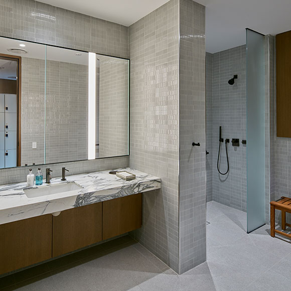 A part of the wellness focused private gym locker room facilities, this stylish bathroom design includes a walk-in accessible shower and a polished marble counter, reflecting contemporary aesthetics. Once again we're balancing warm toned wood finishes with cooler stone and tile for an inviting feel.