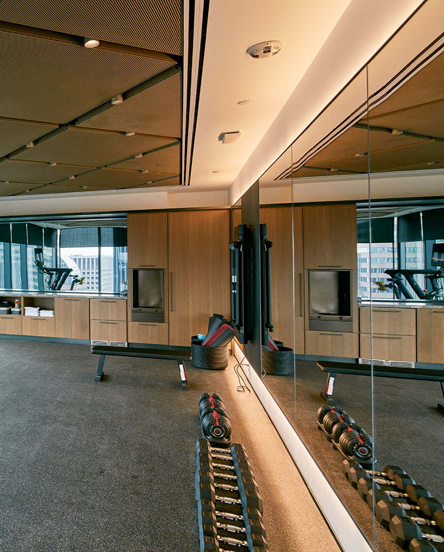 A well-lit luxury gym room adorned with mirrors on the walls and open floor space on the motion-isolated rubber floor provides an expansive feel for exercise and training. This specialized gold metal mesh diamond panel ceiling treatment offers a strong aesthetic and warms the tone of the space, while functionally serving to hide the additional ventilation equipment that keeps the private gym air clean and inviting. The warm wood paneled towel and water station echoes the finishes of the locker rooms.