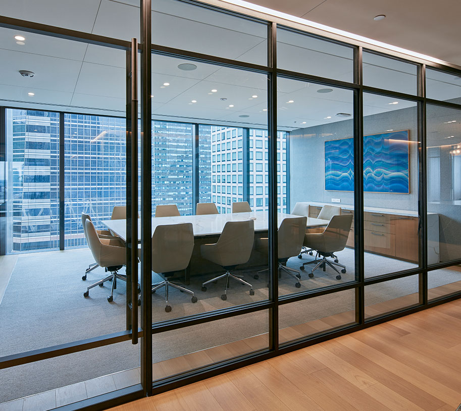 A spacious modern 12-person conference room featuring highly functional acoustic glass partition walls, acoustic fabric and foam wrapped wall panels, and expansive floor-to-ceiling windows that look out into the skyscrapers of Midtown Manhattan's financial hub, allowing natural light to illuminate the space.