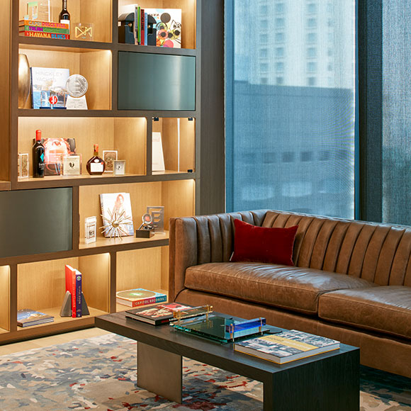 A corner of the welcoming Library meeting room, featuring a plush leather couch, an elegant coffee table, a colorful custom contemporary silk rug, and lit built-in library shelves staged with an array of elegant coffee table books to entertain clients and partners seeking downtime between meetings.