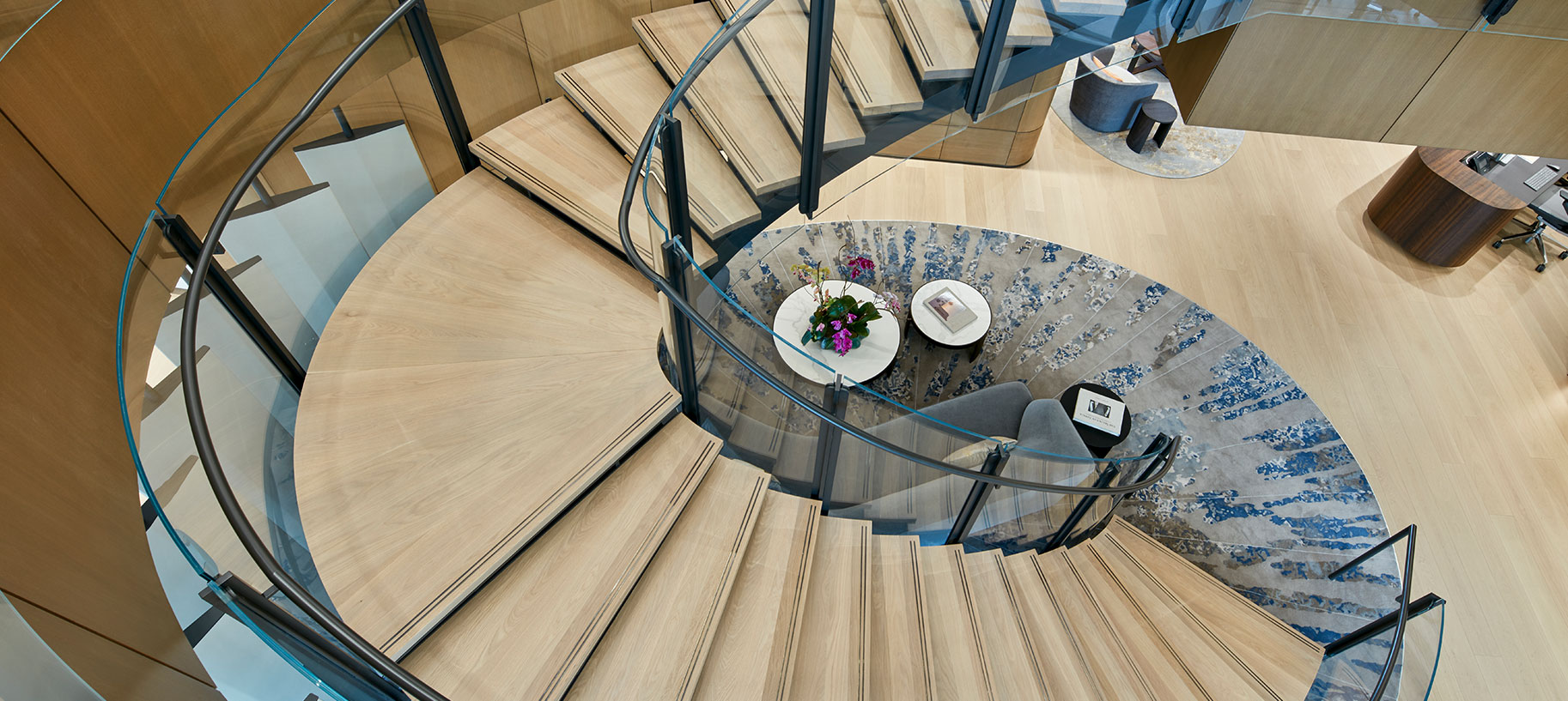 Inside a modern office building, a stylish spiral staircase winds upward, exemplifying innovative architectural design. The custom contemporary oval rug perfectly echoes the staircase opening, and in the left of the image there are operable leather covered acoustic louver panels which can be closed when events are happening in the adjacent flex space, to help prevent noise transferring to the office spaces above.