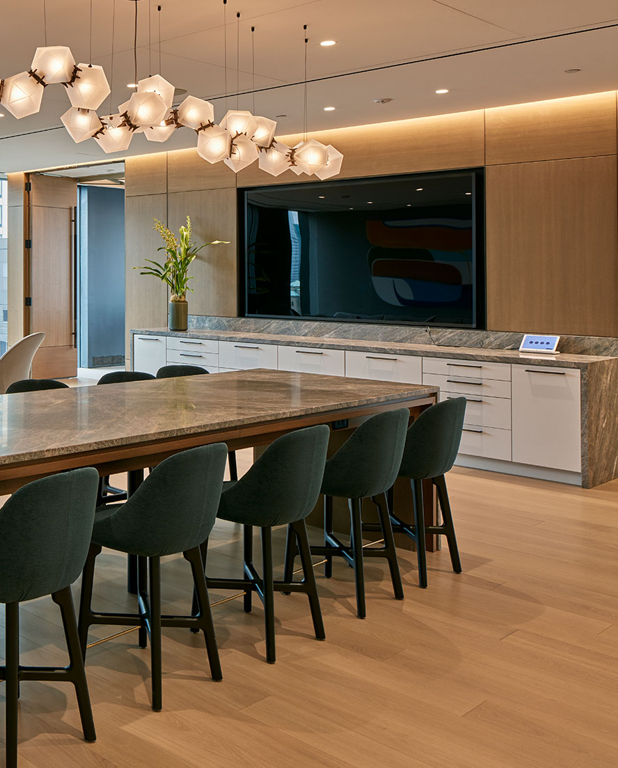 A large breakout collaborative flex space, with a massive island table for lunch, a sleek modern chandelier, and a large built in television for presentations with a functional catering buffet station below