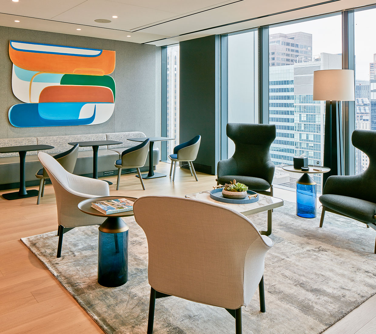 A sleek modern office interior design showcasing a significant painting, part of the all-minority-artists collection, that enhances the workspace's aesthetic appeal. This corner of the communal cafe breakout flex space shows a custom dining bench seat with power for laptops, several bistro tables for individual lunches, and a comfortable lounge area with four chairs overlooking the skyline.