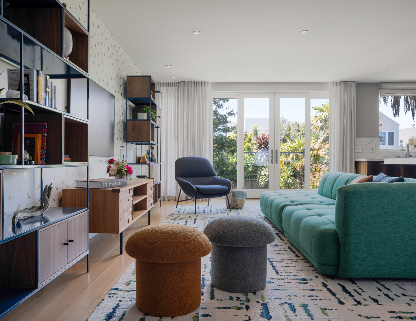 A bright living room showcasing a green couch and a blue chair, with a pair of upholstered mushroom poufs, and wide slider doors perfect for relaxation and inviting gatherings. This modular puffy green sofa is a superstar in the space - it has high style but is still super comfortable for a family friendly interior design.